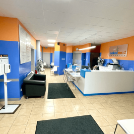 Na zdjęciu widoczna jest poczekalnia dla pacjentów. Ściany są niebiesko-pomarańczowe i kontrastują do białych krzeseł, sufitu i jasnej podłogi. Widać też białą rejestrację.
