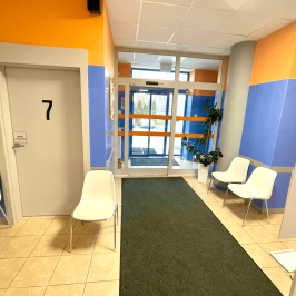 Na zdjęciu widać wejście do poczekalni. Ściany są niebiesko-pomarańczowe i kontrastują do białych krzeseł, sufitu i jasnej podłogi. Zdjęcie wykonane jest od strony poczekalni.