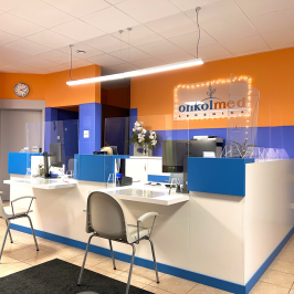 Na zdjęciu widoczna jest biała rejestracja pacjentów z niebieskimi dodatkami. Przy punktach obsługi stoją szare krzesła. Ściany są niebiesko-pomarańczowe i kontrastują do białych krzeseł, sufitu i jasnej podłogi. Po lewej stronie są szare drzi prowadzące do toalet.